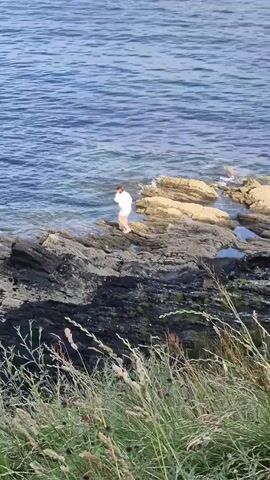 A person in a white shirt and tan shorts is standing on a rocky shore next to the ocean. The water is blue and choppy. There are rocks of various sizes scattered along the shore. In the foreground, there is tall grass and other vegetation. The camera pans slightly to the right, showing more of the ocean and rocks.