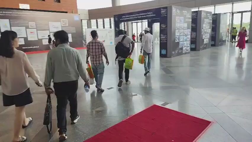 The video shows a wide shot of a busy indoor event space. A red carpet runs diagonally across the foreground. Several people are walking in various directions, some carrying bags. To the left, there are tall, illuminated yellow structures and a black barrier. In the background, a large banner with text and images spans the wall, and further back, there's a stage with people seated and a crowd gathered. More people are milling around throughout the space, with some booths or displays visible. The environment appears to be a convention center or exhibition hall.