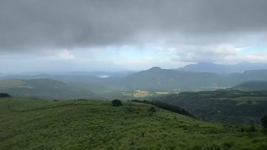 The camera surveys a grassy hill or mountain slope under a cloudy sky. In the distance, a series of rolling hills and mountains are visible, fading into a hazy background. The overall view is of a lush, green landscape, typical of a mountainous region on a cloudy day. The foreground is dominated by the grassy slope, which is uniformly green, suggesting a well-maintained or naturally lush environment. The clouds overhead are dense and gray, indicative of overcast weather. The distant hills are covered in vegetation, further emphasizing the verdant nature of the area. A lake can be discerned in the distance, adding to the geographical detail of the landscape.