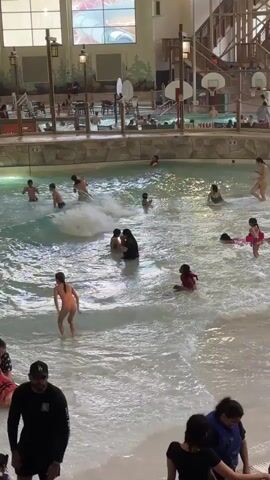 A busy indoor water park wave pool is shown. Numerous people, including adults and children, are in the water, engaging in various activities such as swimming, playing, and riding on inflatable devices. The wave machine is operational, creating moderate waves throughout the pool. In the background, there are water slides, lounging areas, and other park amenities. The overall atmosphere appears lively and fun.