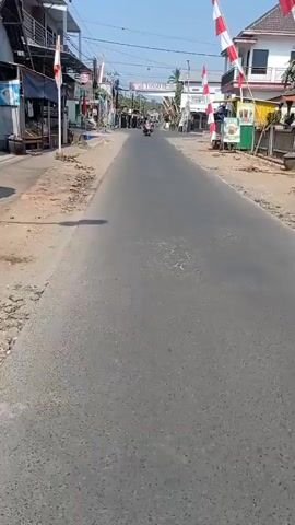 The video shows a wide asphalt road with a cracked surface. The road is lined with buildings on both sides, some of which appear to be shops or residences. Several motorcycles are visible on the road, some moving towards the viewer and others away. Red and white flags are mounted on poles along the roadside, suggesting a local celebration or event. The sky is clear and blue, indicating a sunny day. There are also some trees and utility poles visible in the background. The overall scene depicts a typical street in a town or village, likely in Southeast Asia given the flags and style of buildings.