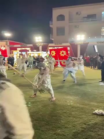 The video shows a group of men performing a dance outdoors at night on a grass lawn. They are dressed in matching silver pants, and most of them are shirtless with painted faces. They hold sticks and move in unison. A man in a white and gold outfit and a red turban stands among them. Many people stand around the perimeter, watching. A cameraman walks through the group, recording the performance. In the background is a building and a large red and white decoration.