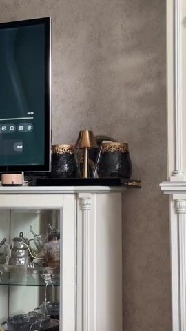 A medium shot shows a shelf on top of a white cabinet. Two black vases with gold trim are on the left and right side of a small golden lamp. The lamp is in the center of the shelf. Behind the lamp is a round mirror. To the left of the shelf is a television. The background is a textured gray wall. Below the shelf is a glass-fronted cabinet with items inside.