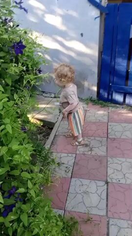 A young child with curly blond hair stands on a tiled patio, facing a large green bush with purple flowers. The child is wearing a tan t-shirt and striped shorts and sandals. The child briefly turns to look at the camera before turning back towards the bush. The scene is outdoors, with a light-colored wall and a blue wooden door visible in the background.