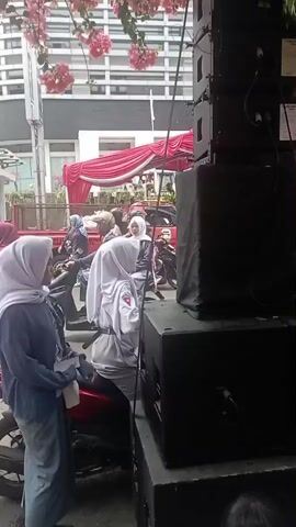 A medium shot shows two young people, possibly students, wearing headscarves. One is on a motorcycle, wearing a white uniform with a red emblem. The other is standing next to the motorcycle, wearing a dark blue top and a light blue headscarf. In the background, there are cars, a red truck with a canopy, and a tall building with many windows. The scene appears to be outdoors, possibly on a street, with pink flowers hanging from above. There are large black speakers stacked on the right side of the frame. The lighting suggests it is daytime.