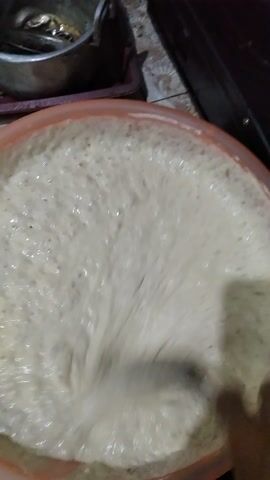 The video shows a close-up view of a cooking process, likely in a home kitchen, given the presence of a stove and what appears to be a tiled backsplash. The scene is dominated by two large containers: one a wide, shallow bowl filled with a light-colored, frothy mixture, and the other a wok or large pan containing hot, bubbling oil.

In the first few seconds, a hand is seen stirring the frothy mixture in the bowl with a utensil. The mixture is very bubbly and appears to be a batter or dough. The camera then shifts to the wok, where pieces of what look like dough are being fried in the hot oil. One piece is already in the oil, puffing up and browning, while another piece is being added or moved around by a spatula. The oil is a deep golden-brown color and is actively sizzling and bubbling around the food.

The lighting suggests it could be daytime, as there are no artificial lights prominently visible, and the overall illumination is relatively natural. The setting appears to be indoors, in a functional, perhaps somewhat utilitarian kitchen space. No specific weather or time of day beyond "daytime" can be inferred.

The focus is entirely on the food preparation. There are no people fully visible, only hands and utensils engaged in the cooking. The activity is clearly the frying of some kind of batter-based food, likely a snack or dessert, given the context of home cooking. The emotions conveyed are neutral, purely observational of the cooking process. There is no visible text within the media.