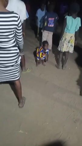The video shows a small child dancing in a sandy area, surrounded by several adults and other children. The child is wearing a striped shirt and is barefoot. The other people in the scene are dressed in casual clothing. The area appears to be outdoors at night. The child is dancing and moving his arms, and music can be heard in the background. The other people are watching the child dance.