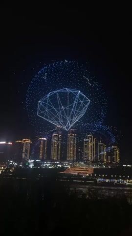A drone show displays a large, geometric diamond shape composed of many lights in the night sky. The diamond is surrounded by a larger, circular formation of scattered lights, also created by drones. Below the drone display, a city skyline is visible, with numerous tall buildings illuminated with interior lights. A river or body of water runs in the foreground, with a dark, silhouetted tree line along its edge. The overall scene is dark, illuminated primarily by the artificial lights of the city and the drone display.