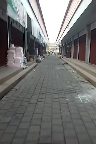 A first-person perspective video shows a person riding a motorcycle down a narrow, paved alleyway lined with closed shops. Stacks of materials, possibly merchandise or construction supplies, are visible outside some of the shops. The sky is bright white, suggesting daylight. A few people are seen in the distance at the end of the alley. The overall atmosphere appears quiet and still, possibly indicating early morning or a time when businesses are not open.