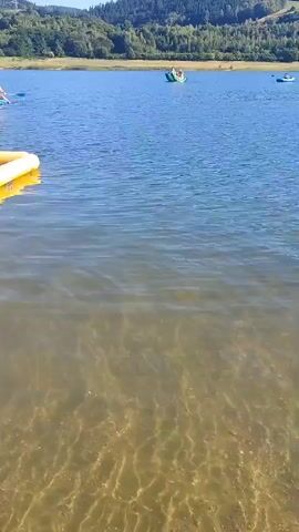 The video shows a body of water with a sandy bottom visible in the foreground. In the distance, there is a shoreline with trees and hills. Several people are visible on the water. On the left side of the frame, there is a green boat with people in it. Further to the right, there are several individuals on paddleboards and some people swimming. A large red inflatable object is also visible in the water. The sun is shining, and the water appears calm.