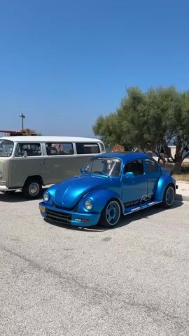 This is a vertical, outdoor video taken on a bright, sunny day with a clear blue sky. The camera pans slowly from right to left, showing a line of vintage Volkswagen vehicles parked on an asphalt surface. In the foreground, a bright blue Volkswagen Beetle with "RS" graphics on the side is prominently displayed. To its left, a light green Volkswagen Type 2 van, also known as a Kombi or Bus, is parked, with surfboards on its roof rack. Further down the line, more Volkswagen vehicles, including several more Type 2 vans in various colors (red, white, grey, yellow, green) and a few other classic car models, are visible. The background features sparse vegetation, some trees, and a distant view of the sea. The scene appears to be a gathering or exhibition of vintage Volkswagen vehicles, possibly at a seaside location. There are no people clearly visible in the video.