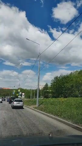 The video shows a view from inside a vehicle driving on a paved road. The sky is blue with large, white, fluffy clouds. Trees and green grass line the right side of the road. There are several cars on the road ahead, some of which appear to be stopped or moving slowly in traffic. There are also streetlights and traffic signals visible along the road. The sun is shining, casting shadows.