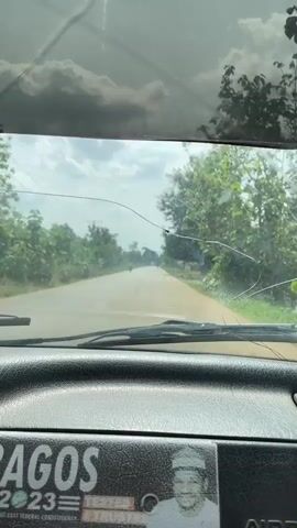 From the perspective of a vehicle's dashboard, the view is through a cracked windshield onto a paved road. A motorcyclist is visible in the distance on the road. Trees line the sides of the road under a cloudy sky. The vehicle's wipers are visible and stationary. The dashboard includes a sticker with an image of a person on it.