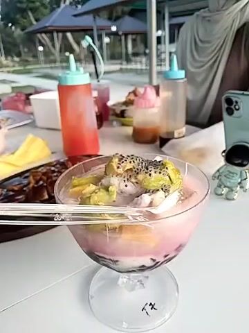 The scene is an outdoor setting where a woman with her head covered, possibly a hijab, is seated at a white table. On the table, there is a dessert in a glass footed bowl that contains layers of pink liquid, dark jelly balls, chunks of fruit, and black seeds sprinkled on top. There are also bottles of red and dark brown liquids, a plate of food, and a small astronaut figurine on the table. The woman is not directly interacting with the dessert, and the focus is on the dessert itself. In the background, there is a gazebo-like structure, trees, and other greenery. The lighting suggests that it is daytime.