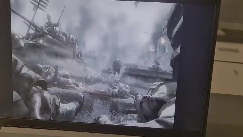 The video displays a black and white still image of a battlefield. In the foreground, a person's arm and leg are visible, appearing to be a dead soldier. In the mid-ground, numerous fallen soldiers are scattered across a muddy, debris-strewn area. Bloodstains are visible on the ground. In the background, a bombed-out city is depicted, with smoke billowing into the air. Several birds are flying overhead, and what appear to be anti-aircraft guns are visible on the left side of the image. The overall scene suggests the aftermath of a fierce battle, with a grim and chaotic atmosphere.