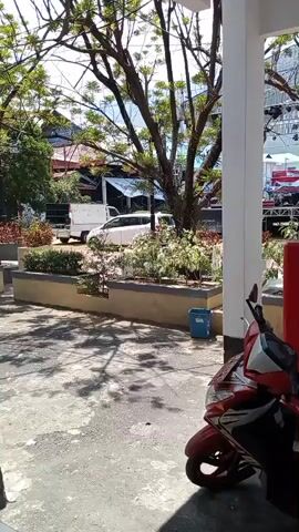 A red and white motorcycle is parked on a paved area. Beyond the motorcycle, there is a raised planter box with decorative plants. Further in the background, a white van and a white truck are parked. There is a stage structure with lights and sound equipment set up in the distance. A large red generator is also visible. The scene appears to be outdoors, possibly in front of a building, with trees and a mountainous landscape in the far background.