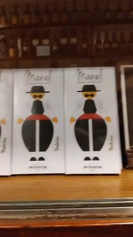 The camera pans across a row of chocolate boxes displayed on a wooden shelf. The boxes have different designs depicting traditional Portuguese figures and motifs. Behind the shelf, a wall is lined with bottles, likely in a bar or shop setting.
