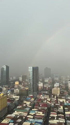 This is a vertical, eye-level drone shot looking down on a dense urban cityscape under a hazy, overcast sky. A faint rainbow arches across the upper portion of the frame, extending from the left side towards the right. The city is characterized by a mix of modern high-rise buildings and older, lower-rise structures with colorful, tin roofs. The buildings are tightly packed, with streets and alleyways visible between them. The overall atmosphere is muted, suggesting a recent rain or ongoing dampness in the air. No people or vehicles are discernible in the shot due to the distance and haze.