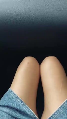 The video opens with a close-up view of a person's knees and the top of their denim jeans, suggesting they are sitting in a vehicle. The camera then pans up, revealing the interior of a car's dashboard and windshield. Through the windshield, a road is visible, lined with trees and some buildings. The scene appears to be a rural or suburban setting, possibly on an overcast day, as the sky is gray. The video then continues to show the road ahead as the vehicle is in motion, passing by more trees and vegetation.