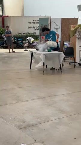 A young person wearing goggles and orange gloves pours a white, gaseous substance from a large metal cylinder into a blue bucket on a table covered with a white sheet. The substance is producing a visible white vapor. The person is standing in an outdoor or semi-outdoor space with a paved floor. In the background, there is a wall with writing and several strollers. Another person stands to the left, observing.