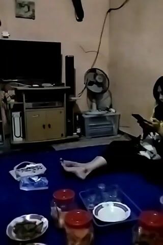 The video shows a dimly lit room with a blue rug. In the foreground, there are several plates and containers of food, as well as some glasses. A person's bare feet are visible in the lower right corner. In the middle ground, there are two people sitting on the floor, looking at their phones. To the left, a person stands and walks away from the camera. In the background, there is a television set on a stand, a fan, and shelves filled with items. The overall atmosphere appears casual.