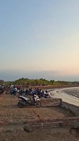 The video begins with a wide shot of a coastline at sunset. The sky is a gradient of pale blue and soft orange, indicating the sun is low on the horizon. The water appears calm, with a muddy, textured surface suggesting low tide. Along the shore, there's a line of trees, and a few people are visible sitting by the water or on a paved area. Several motorcycles are parked nearby. As the video progresses, the camera focuses on a dilapidated brick structure with graffiti on its wall. A young person is seen sitting on a low wall in front of the structure, looking out towards the water. The sun, a bright orange orb, is clearly visible in the sky, casting a warm reflection on the water's surface. The scene is peaceful and contemplative, capturing the serene beauty of a coastal sunset.