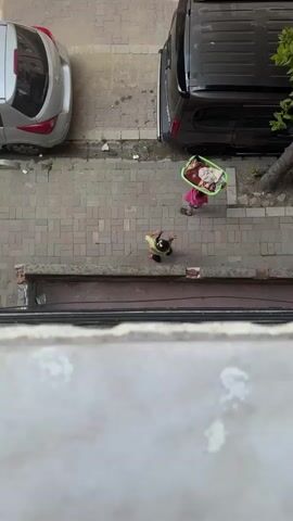 Two people are walking on a sidewalk. One person is carrying a green basket filled with items on their head. Another person is walking behind them. A third person is visible further down the sidewalk, appearing to be doing some kind of exercise on the ground. A black van is parked on the street next to the sidewalk, and a silver car is parked further up the street. There is a tree on the sidewalk next to the van. The scene appears to be outdoors in an urban environment, with a building or wall in the foreground.