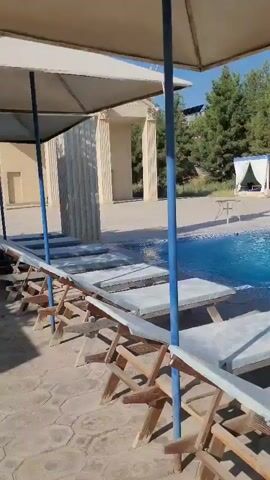 A video shows a swimming pool area. Several lounge chairs are lined up along the edge of the pool. There are umbrellas providing shade over some of the lounge chairs. In the background, there is a building with columns, trees, and cabanas. A round table with chairs is also visible near the pool. The water in the pool is blue and appears to be clean. The sun is shining, indicating it is daytime. There are no people visible in the video.