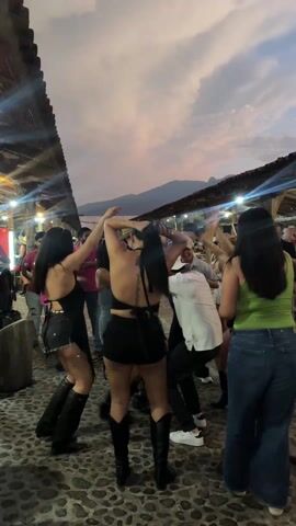 A vibrant outdoor gathering in Amatitán, Mexico, unfolds at dusk, with people actively dancing to live music on a cobblestone surface. In the foreground, three women are seen from behind, energetically moving. The woman on the left wears black shorts, a black top, and tall black cowboy boots, with her arms raised. Central to the frame, another woman, also in black shorts and a black halter-style top, with her dark hair tied up, mirrors the raised arm gesture, also sporting tall black boots. To the right, a woman in a green tank top and blue jeans sways rhythmically.

Behind these individuals, a diverse crowd of people is visible, some dancing, others socializing. A musician wearing a pink shirt and holding a saxophone can be seen on the far left, suggesting a band is performing. Several individuals in the background are wearing cowboy hats, contributing to a festive, possibly Western-themed, atmosphere.

The scene is illuminated by string lights strung across rustic, wooden overhead structures, possibly part of a covered walkway or an open-air market. The sky above displays the soft, fading colors of sunset, with hues of pink, orange, and grey clouds, and distant mountains are silhouetted against the horizon. The overall impression is one of lively celebration and enjoyment in a traditional setting.