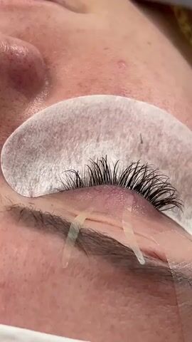 A close-up shot focuses on an eye as a person applies eyelash extensions. Tweezers hold a single, thin extension, which is being carefully placed onto a natural lash. A white under-eye patch is visible beneath the lashes, and strips of tape secure the lower lashes. The skin of the person's face is visible around the eye, and another person's hand is partially visible holding the eye open.