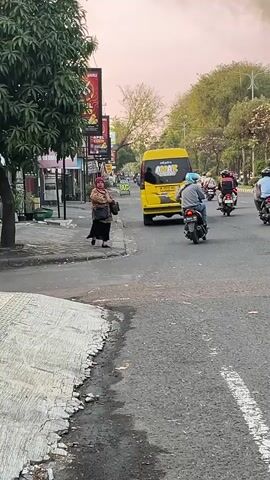 The video opens with a person standing on the sidewalk, dressed in a hijab and traditional clothing, carrying a bag. They are near a street with several motorbikes passing by and a yellow van in the distance. The street is paved with asphalt and has a white line marking the lanes. Trees and buildings line the sides of the street, and the sky is visible in the background. The scene appears to be an urban or suburban environment during the daytime, possibly late afternoon due to the lighting. The person on the sidewalk seems to be observing the traffic. The motorbikes are moving in the same direction as the van, and their riders are mostly wearing helmets.