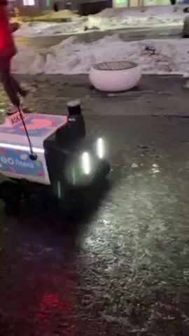 A delivery robot rolls across a wet, paved area at night. The robot is white and black, with a red flag on a pole. There are lights on the front and back of the robot. The robot has the logo for “Lavka” on its side. The area around the robot is wet and reflective. In the background, there are buildings, streetlights, snow, and people walking. A person with purple hair walks past the robot on the left side of the frame.