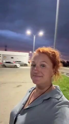 A woman with red hair smiles at the camera while standing outside at dusk. She is wearing a light blue shirt. Behind her, there are streetlights, a sidewalk, grass, and a road with vehicles. In the final frames, a man appears and waves at the camera.