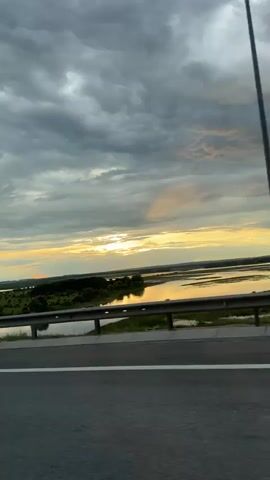 This media file captures a scenic view from a bridge, likely a highway overpass, looking out over a wide expanse of water and a tree-lined landscape. The time of day appears to be dusk, with the sun setting on the horizon. The sky is filled with dramatic, heavy clouds, tinged with yellow and orange hues from the setting sun, creating a sense of twilight and perhaps impending weather change. The water reflects the colors of the sky, adding to the ethereal quality of the scene.

The foreground features the edge of the bridge, with a metal guardrail and a paved road surface visible. Beyond the guardrail, a strip of green land with scattered trees leads to the expansive body of water. The overall impression is one of natural beauty and tranquility, with the vastness of the sky and water suggesting a sense of open space. There are no people or specific objects that stand out, emphasizing the natural landscape. The location context of Gavrilkovo, Russia, places this scene in a rural or semi-rural environment, consistent with the wide-open, natural vista presented. The mood is one of peaceful observation, perhaps during a journey. No text is visible in the media file.
