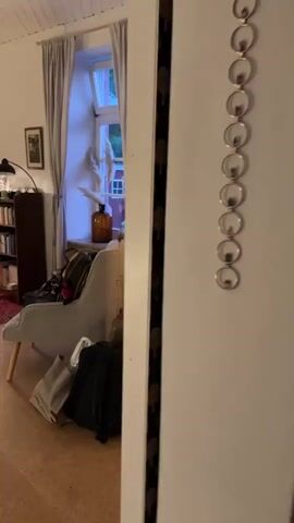 The video opens showing a glimpse of a white door frame with a black and white patterned ribbon running vertically down it. The camera then pans left, revealing a room with several pieces of luggage near a white wardrobe. To the left of the wardrobe is a window with grey curtains, and a stuffed armchair is positioned in front of it, with bags on and around it. The camera then moves further left to show a bookshelf filled with books, a floor lamp with a black shade, and two decorative palm tree lamps on a red patterned rug. The walls are white, and the ceiling appears to be made of white wood planks. The lighting in the room is dim, with some areas illuminated by the lamps and the light coming through the window.
