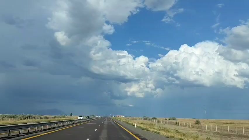 A first-person perspective video shows a car traveling on a highway under a partly cloudy sky. The road is asphalt with yellow and white lines. On the left side of the road, there is a metal barrier and then a field with trees. On the right side, there is a fenced field with dry grass and scattered trees. The sky is a mix of dark, stormy clouds and bright blue sky with white fluffy clouds. Several cars are visible on the highway in the distance. There are utility poles on the right side of the road. The video appears to be taken on a sunny day, possibly before or after a storm, given the presence of both dark and bright clouds.