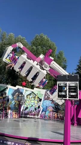 The video shows a carnival ride called "Move It" in motion on a sunny day. The ride is primarily pink and white, with several arms that swing and rotate, lifting gondolas of people into the air. The gondolas themselves also spin. The ride is decorated with colorful graphics, including images of animated characters. A large Ferris wheel can be seen in the background, along with green trees and a bright blue sky. There is a platform at the base of the ride where people are visible standing. The ride appears to be operating at a moderate pace, with people on the ride looking excited.