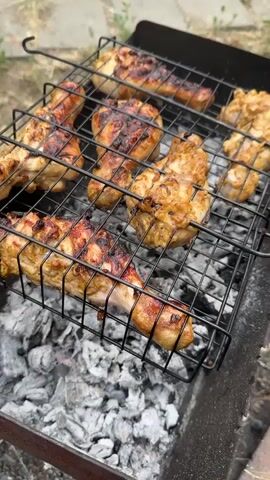 The video shows a close-up of chicken pieces grilling over hot coals in a barbecue. The chicken legs are arranged on a black metal grill basket, with several pieces visible. They appear to be seasoned and are browning nicely with visible grill marks. The coals underneath are glowing and producing heat, indicated by the shimmering air above them. The background is slightly blurred but suggests an outdoor setting with dry grass or dirt. The lighting suggests it might be daytime, perhaps late afternoon or early evening, given the natural light. No people are visible in the frame. The overall impression is of food being prepared for a meal outdoors. There is no visible text in the video. The location context provided is Biysk, Russia.