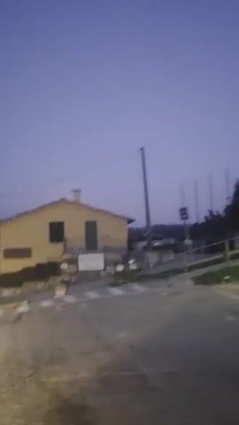 The video shows a yellow house with a red tile roof in what appears to be a rural setting. There are a few signs visible: a triangular sign with two figures walking, indicating a pedestrian crossing, and another triangular sign with two figures walking. A street light is also visible. The sky is a deep twilight blue, suggesting it is either dawn or dusk. The ground is paved with what looks like gravel or dirt. The house has two windows and a door on the side visible, and a balcony above the door. There's a fence in front of the house made of stone and metal. The overall mood is quiet and still, perhaps peaceful or slightly eerie due to the low light. The location is identified as Fumane, Italy, which implies an Italian architectural style for the house and potentially the road. There is no discernible activity beyond the slow movement of the camera, which seems to be moving along a road.