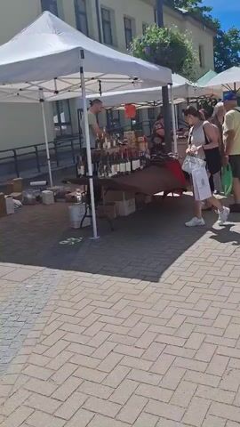 The video shows a market scene on a sunny day. In the foreground, a person pushes a stroller. In the mid-ground, a white tent is set up, with a vendor standing behind a table displaying bottles of wine. A woman is looking at the bottles. Several other people are walking around the market, browsing stalls under white tents. The ground is paved with brick. Trees and buildings are visible in the background.