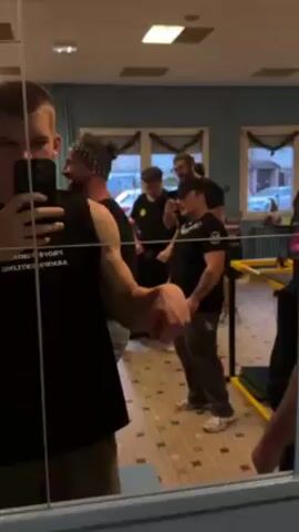 Here's a detailed description of the video:

A young man stands in front of a mirror, holding a smartphone to record a video. He's wearing a black sleeveless shirt with white lettering. He flexes his arm, showing off his bicep. In the background, reflected in the mirror, are several other people in what appears to be an indoor recreational space. Some individuals are standing and talking, while one person gestures with their hand. The room has blue walls with decorative trim, and windows with visible street scenes outside. The floor has a tiled pattern. There is a table with a green cushion. The lighting in the room is bright and even.