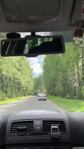 The video shows the interior of a car driving on a road. The road is paved and appears to be surrounded by trees on both sides. In front of the car, there are two other vehicles. The car's dashboard is visible, along with the rearview mirror. A small air freshener hangs from the mirror. There is also a mobile phone holder on the dashboard with the brand "WALKER" visible. The sky is visible in the distance, appearing partly cloudy.