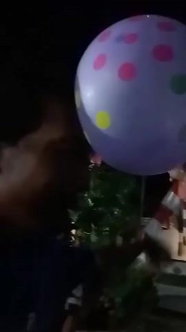 A group of people are at a party. A large, purple polka-dotted balloon is being held up by someone. The camera is low and shaky, capturing the action from a close-up perspective. It is nighttime and the background is dark with some lights visible.