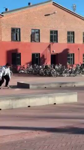 The video shows a person skateboarding on a gray concrete ledge in an outdoor setting. The skateboarder is wearing a light-colored hooded sweatshirt, dark pants, and a dark hat. They are balancing on the ledge, with their arms extended for balance, and then turn slightly, raising one arm in the air. The person then attempts a trick, the skateboard leaving the ledge, and the person lands back on the skateboard. The surrounding environment includes a red brick building with dark windows and a dark door, a large number of parked bicycles behind the ledge, and a red brick-patterned ground. The sky is clear and blue. Other people are visible in the background.