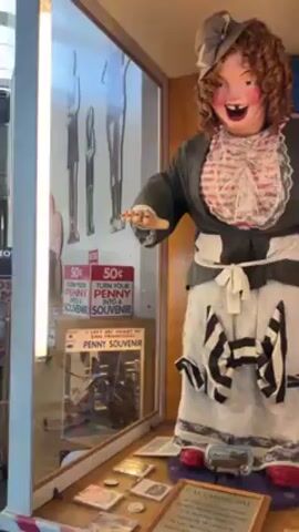 The video shows a Laughing Sal animatronic figure in a display case inside an arcade or museum. The figure is wearing a gray jacket over a white blouse with lace detailing, a white skirt with black and white striped accents, and a gray bonnet. A sign below the figure reads "Laughing Sal." Beside the figure is a coin-operated machine that creates penny souvenirs. The camera then pans up to reveal an arcade or museum filled with vintage arcade games and decorations. Several people can be seen walking around and playing the games. The space has a high ceiling with exposed metal beams and supports. Banners and signage are hanging from the ceiling.