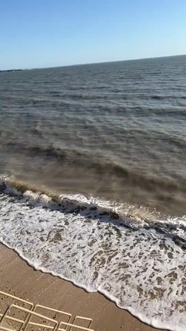The video shows a shoreline with waves gently lapping against the sand. The water is a murky brown color and appears somewhat choppy. The sky is clear and blue, with the sun shining brightly, creating a glare on the water's surface in the distance. In the far distance, a faint silhouette of a boat or ship can be seen. The foreground of the video shows a portion of a white, wire-mesh fence or barrier on the sandy beach.