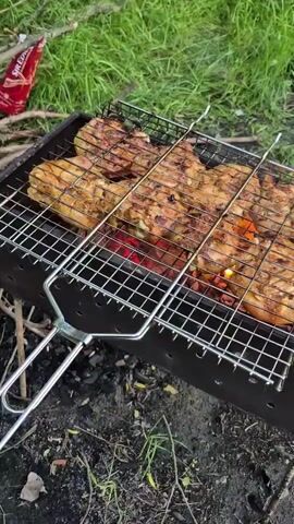 The video shows chicken pieces cooking on a grill over a fire. The grill is made of metal mesh and has a handle. The chicken is seasoned and appears to be browning. The grill is placed on top of a metal barbecue pit filled with burning coals. In the background, there is green grass and some branches. A red bag is also visible in the upper left corner. The scene suggests an outdoor cooking activity.