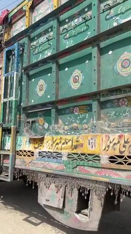 A camera pans right to left, revealing a highly decorated truck parked on a dusty, unpaved road under a clear sky. The truck's cab and a portion of its cargo area are visible, featuring intricate, colorful patterns and designs in red, green, blue, yellow, and brown, with small, decorative metal chains hanging from the bottom edge of the cab. The truck's front wheel is visible on the left. In the background, a wooden utility pole leans over a pile of rubble. Further back, brick buildings, partially constructed structures, and more dusty terrain are visible, suggesting a rural or semi-rural setting. A power line extends from the top of the utility pole towards the right, out of frame.