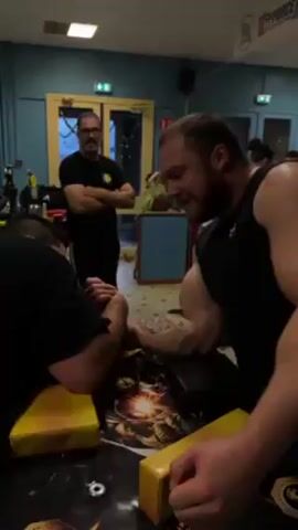 The video shows an indoor arm wrestling match in progress. A man in a black tank top is seen exerting himself as he engages in the competition. Other arm wrestlers can be seen at nearby tables. Several spectators watch the matches, including one man with a beard and glasses standing with his arms crossed. The room is decorated with blue and white walls, and there are tables and chairs scattered around. Banners are visible along the ceiling.