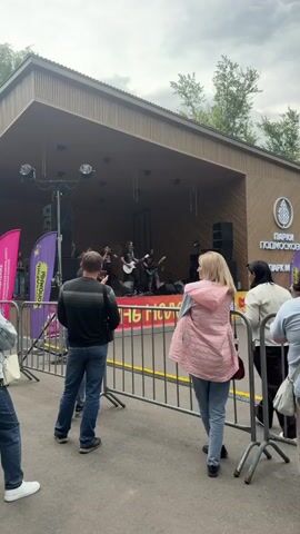 A band performs on an outdoor stage. A crowd watches from behind a metal barrier. The stage is covered by a wooden awning and decorated with banners. Trees are visible in the background. A woman in a pink jacket stands facing the stage, her back to the camera. A man in a black jacket and jeans holds up a phone to record the performance. Two women stand to the left of the man, looking towards the stage. The overall environment appears to be a park or outdoor event space.